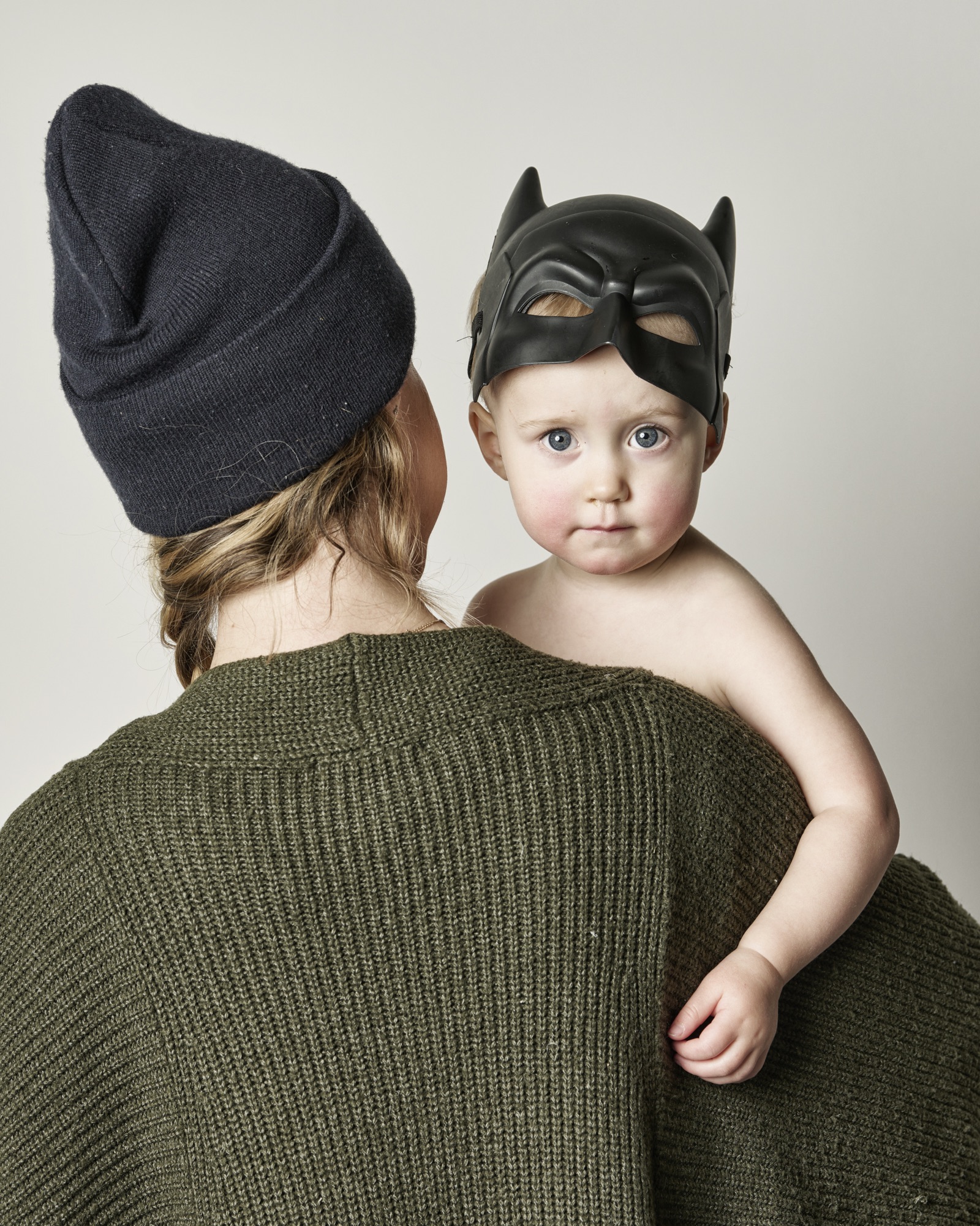 Toddler in a bat mask held by a parent, family portrait by Winky Lewis